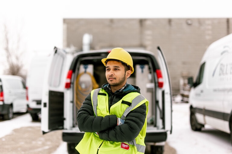A service worker in a reflective safety vest and yellow hard hat stands confidently with arms crossed in front of a van, representing the reliable and professional workforce that benefits from van upfitting. The backdrop features a snowy parking area and company vans, emphasizing a work environment that values safety and efficiency.