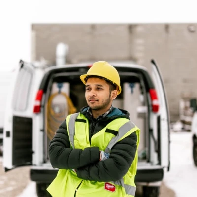 A service worker in a reflective safety vest and yellow hard hat stands confidently with arms crossed in front of a van, representing the reliable and professional workforce that benefits from van upfitting. The backdrop features a snowy parking area and company vans, emphasizing a work environment that values safety and efficiency.