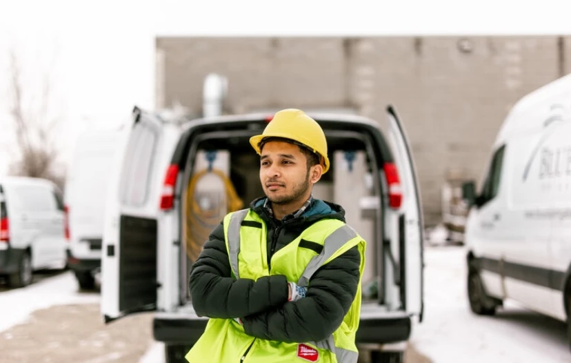 A service worker in a reflective safety vest and yellow hard hat stands confidently with arms crossed in front of a van, representing the reliable and professional workforce that benefits from van upfitting. The backdrop features a snowy parking area and company vans, emphasizing a work environment that values safety and efficiency.