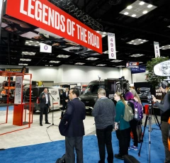 Attendees at a trade show with a large overhead sign reading 'LEGENDS OF THE ROAD'. The exhibit features various vehicles and booths, with event-goers engaging in conversation and exploring the displays.
